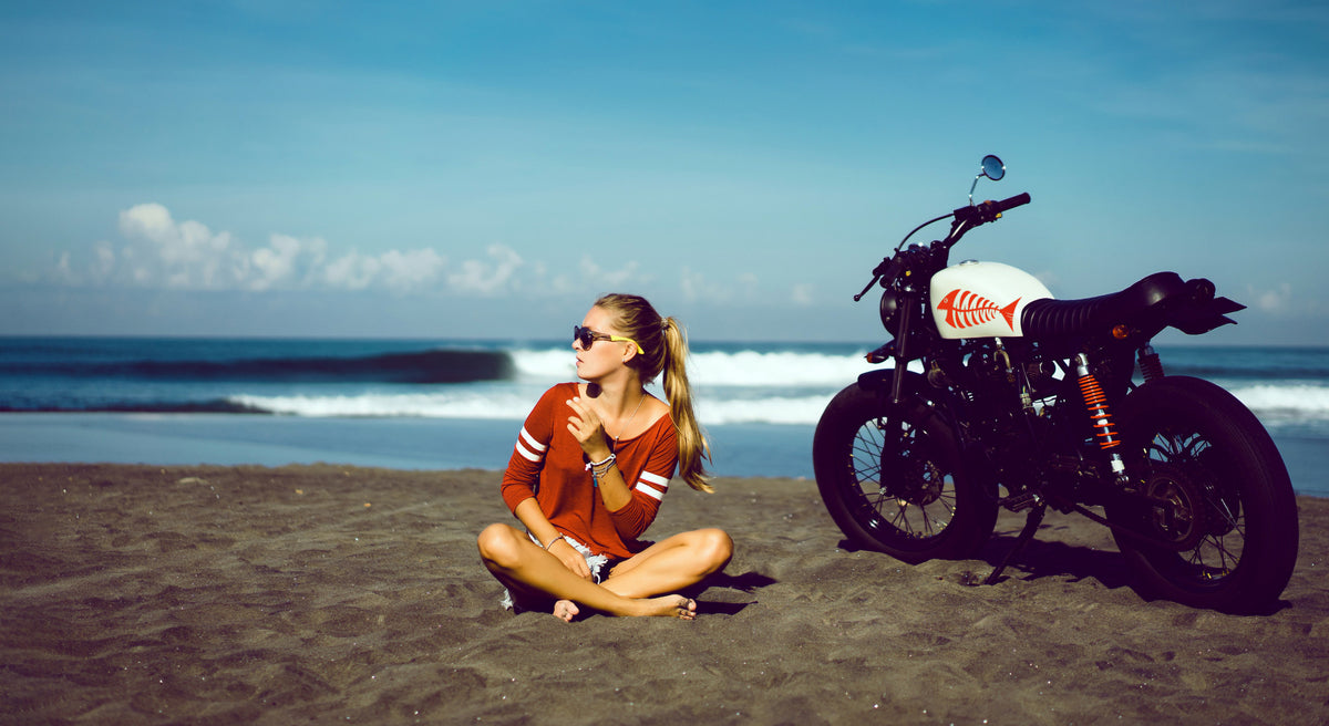 Frau sitzend am Strand. Motorrad mit Aufkleber im Hintergrund